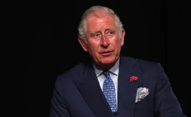 King Charles III in a navy suit with a patterned tie, looking slightly to the side against a dark background.