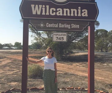 A woman stands by a sign reading "Wilcannia, Central Darling Shire, Population 745" under a sunny sky.