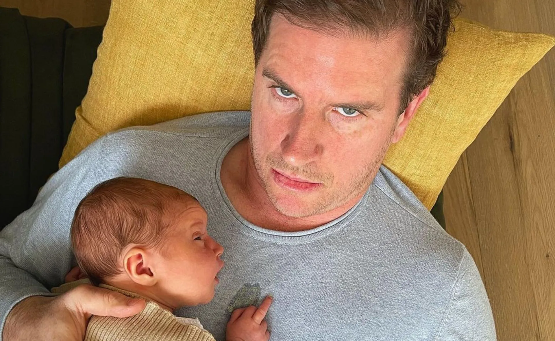 A man with a serious expression holds a sleeping newborn on his chest, laying on a yellow pillow.