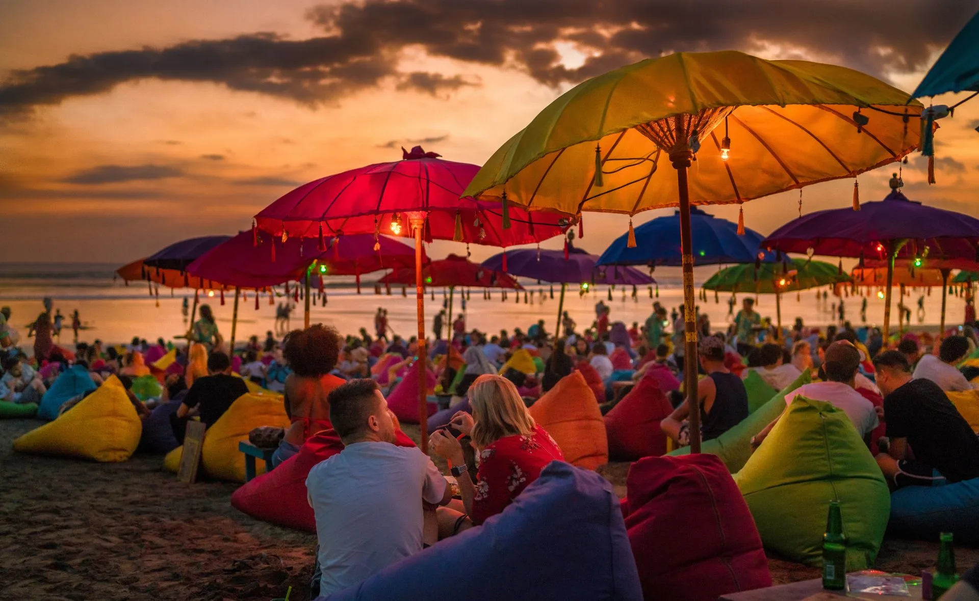 Crowded beach scene at sunset with colorful umbrellas and beanbags, people relaxing and socializing.