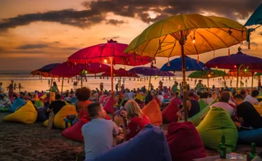 Crowded beach scene at sunset with colorful umbrellas and beanbags, people relaxing and socializing.