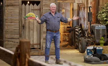 A man stands with open arms in a rustic setting, beside a wooden shed and an old tractor, with a dog resting nearby.