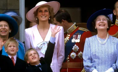 Royal family smiling and looking up, child waving; Princess Diana in pink, Queen in blue hat.