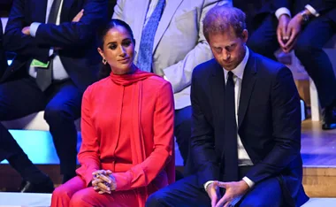 A woman in a bright red outfit sits beside a man in a dark suit, both in a formal setting.