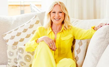 Blonde woman in a bright yellow outfit sits on a plush couch, smiling at the camera.