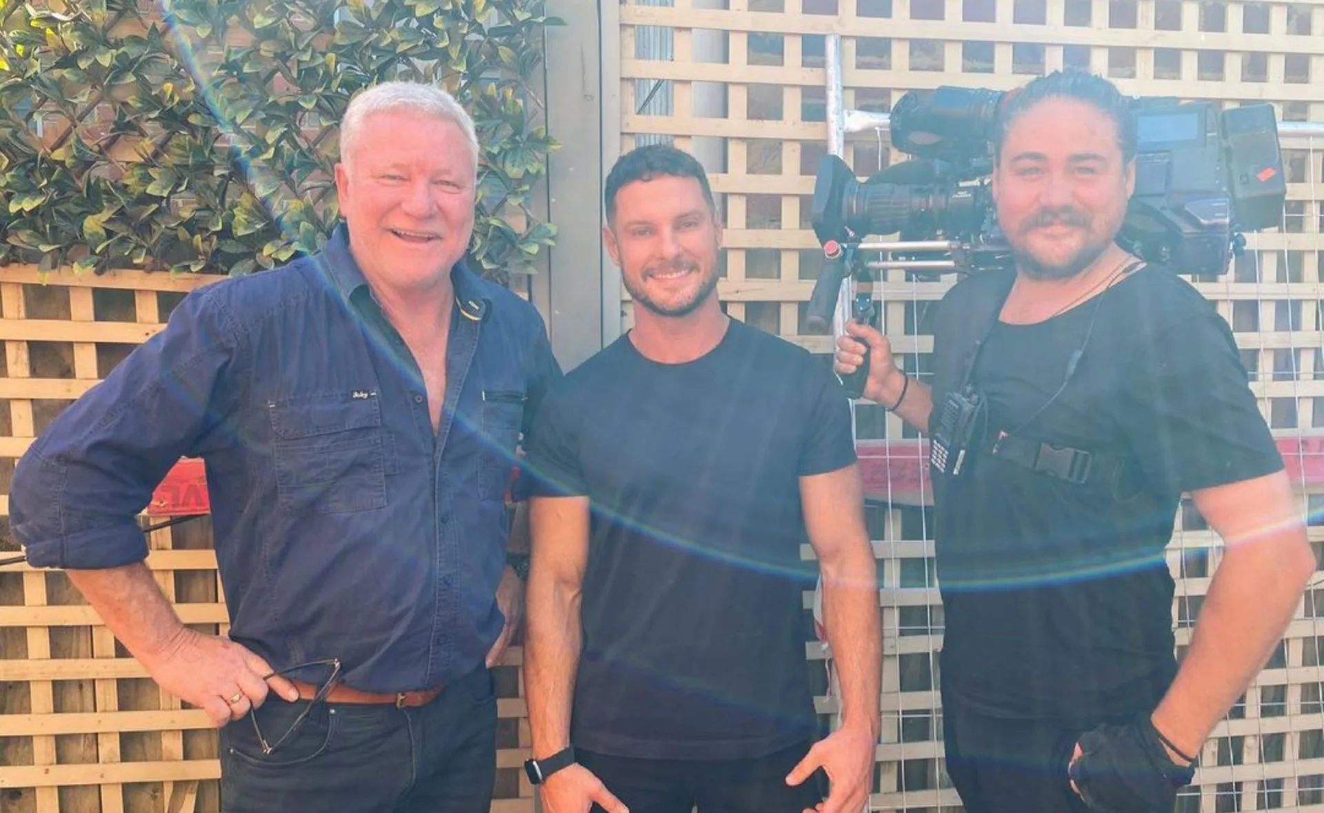 Three men smiling, one holding a TV camera, standing outdoors in front of a lattice fence with greenery.