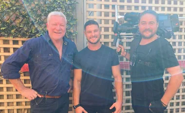 Three men smiling, one holding a TV camera, standing outdoors in front of a lattice fence with greenery.