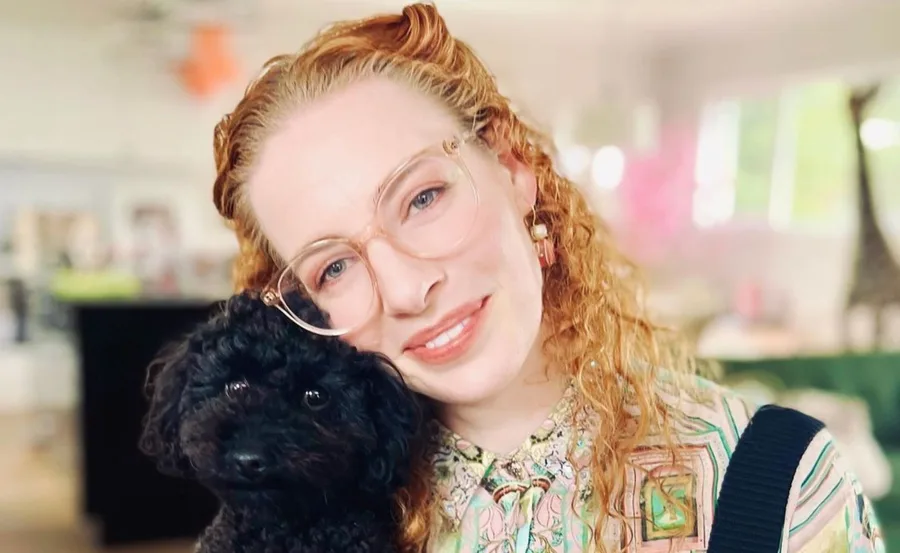 Woman with curly red hair and glasses smiling, holding a small black dog indoors.