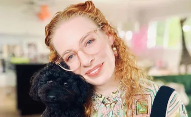 Woman with curly red hair and glasses smiling, holding a small black dog indoors.