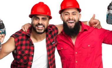 Two men wearing red construction helmets, smiling and holding power tools, with arms around each other in a friendly pose.