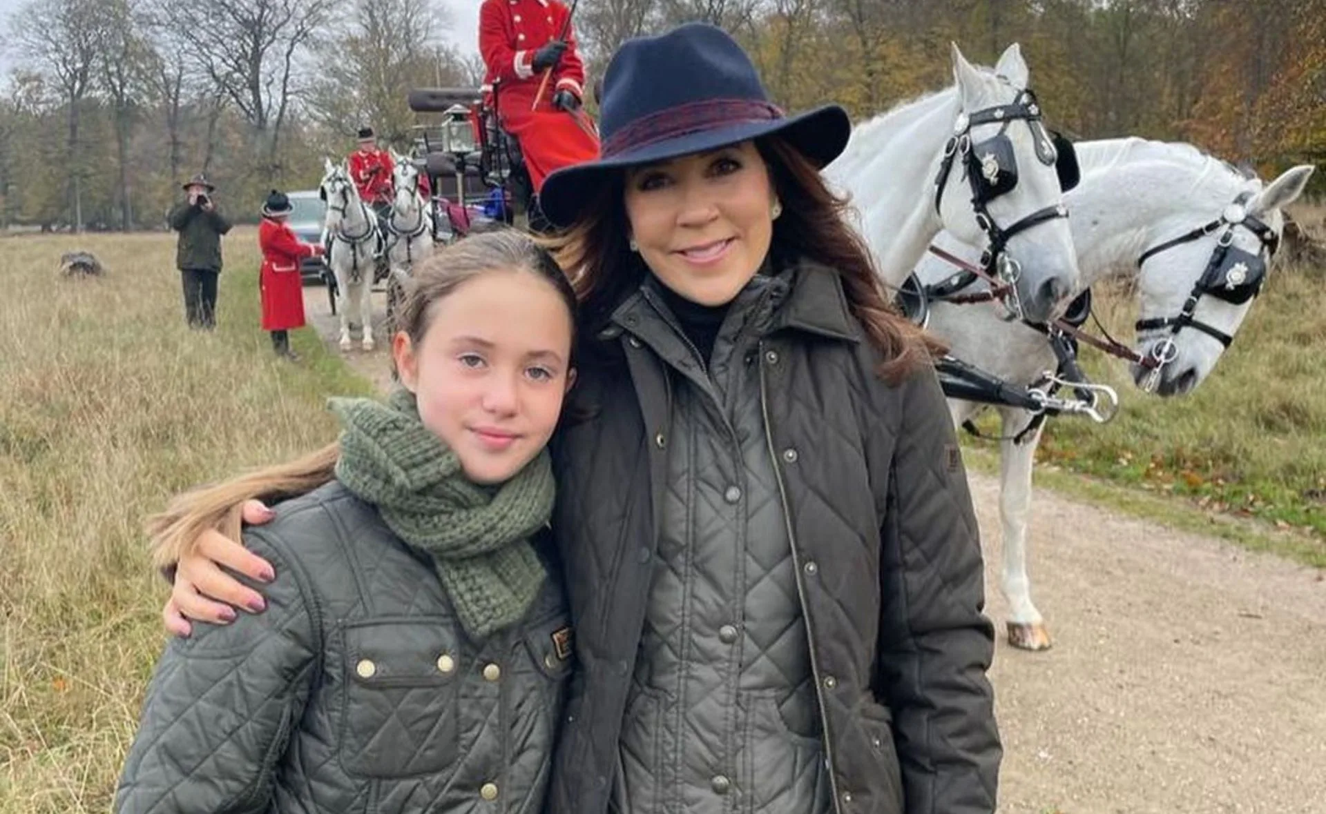 Mother and daughter in matching coats, posing near white horses and a carriage outdoors.