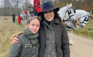 Mother and daughter in matching coats, posing near white horses and a carriage outdoors.