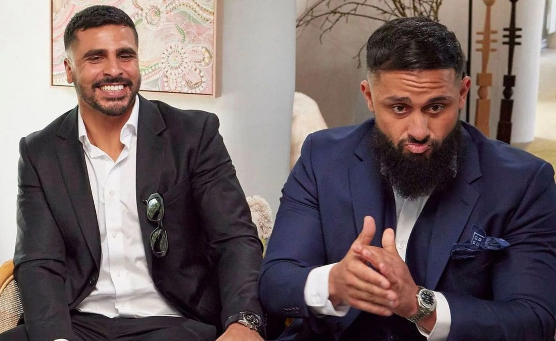Two men in suits sitting indoors, one smiling and the other clasping hands, with artwork and decor in the background.