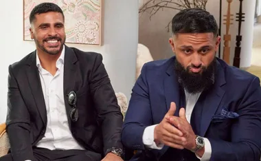 Two men in suits sitting indoors, one smiling and the other clasping hands, with artwork and decor in the background.