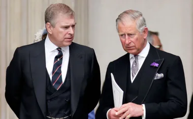 Two men in formal suits conversing, one holding a document with a flower in his lapel.