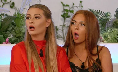 Two women on Love Island, with surprised expressions, sitting outdoors amidst greenery.