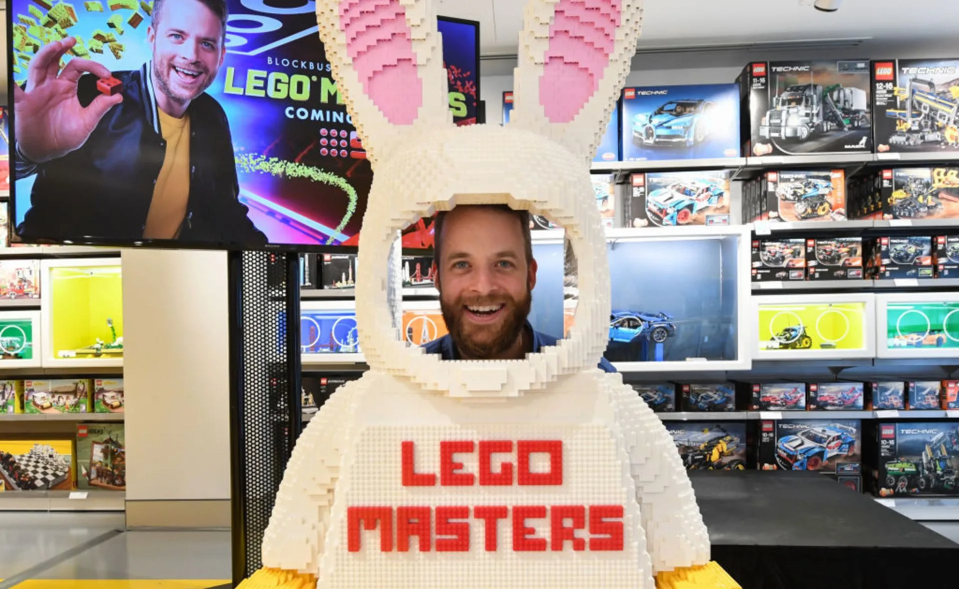 Person smiling through a LEGO bunny suit with "LEGO Masters" text, surrounded by LEGO sets and a promotional screen.