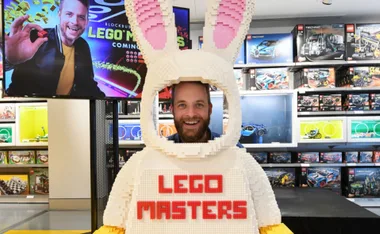 Person smiling through a LEGO bunny suit with "LEGO Masters" text, surrounded by LEGO sets and a promotional screen.