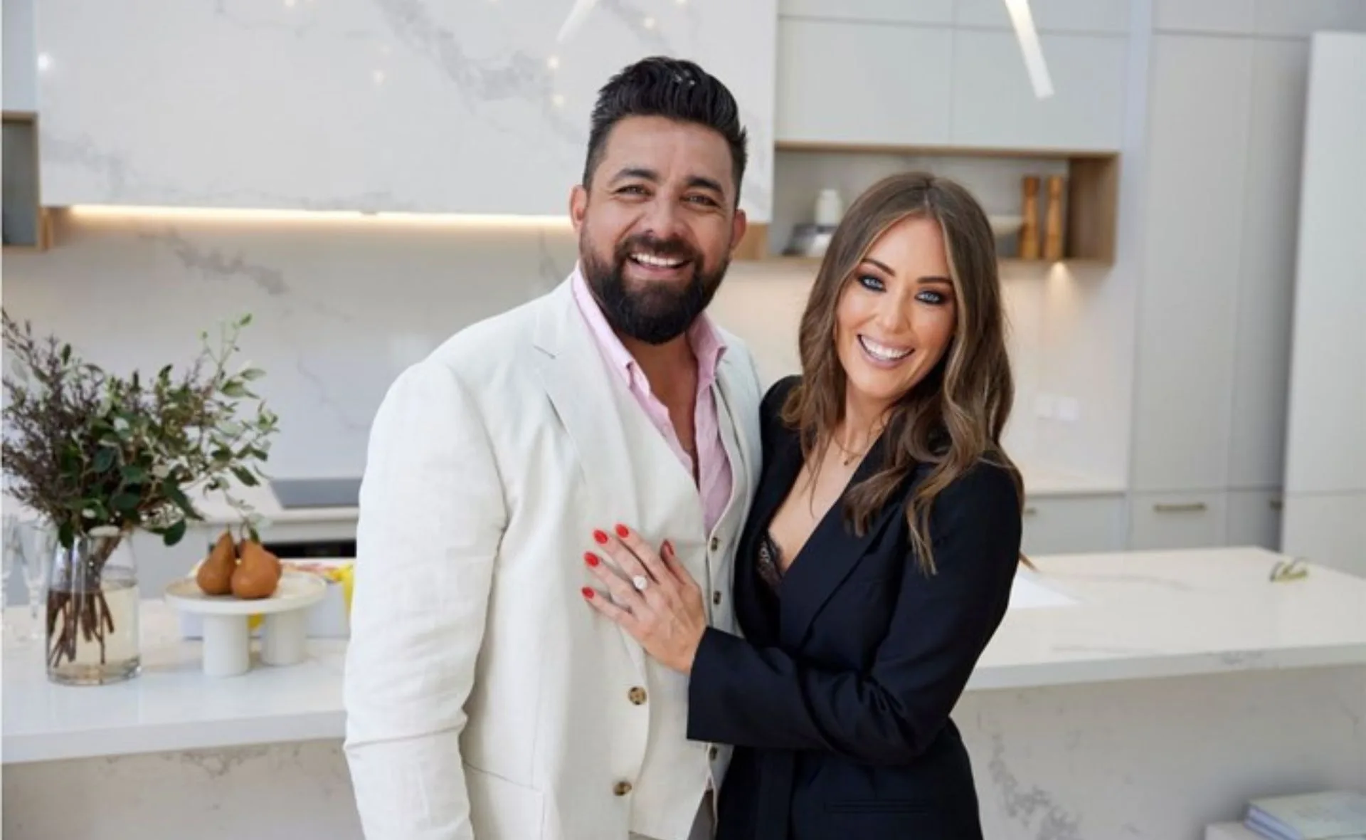A smiling couple in formal attire stands in a modern kitchen setting with a vase and fruit on the counter.