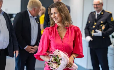 A woman in a bright pink blouse holding a bouquet, smiling, with people in the background.