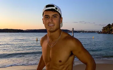 Shirtless man on the beach at sunset, wearing a backwards cap, with water and distant hills in the background.