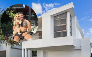 Man with a tattoo holding a dog, sitting outdoors in a garden. Modern white house under a blue sky in the background.