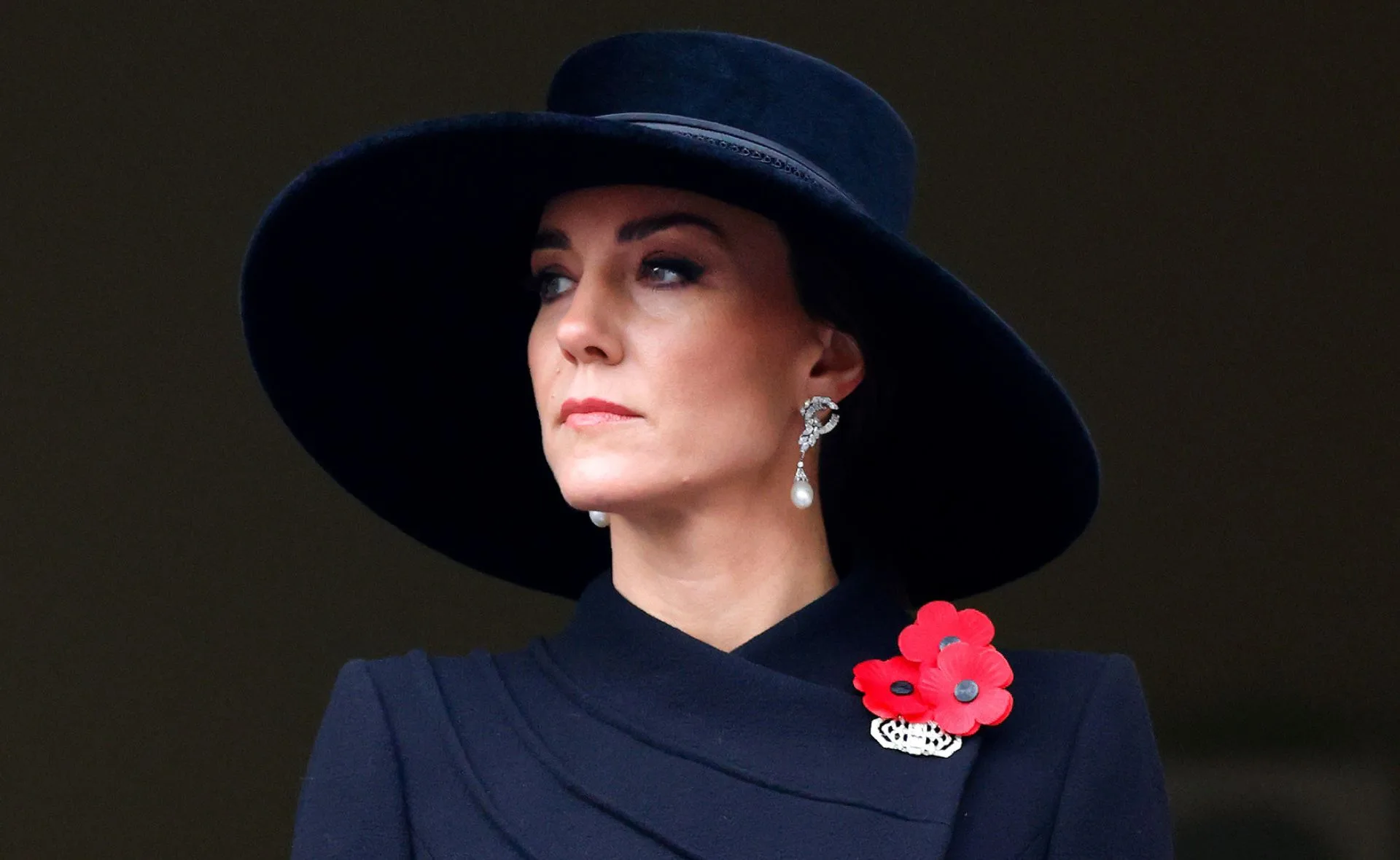A person wearing a wide-brimmed black hat and pearl earrings, with red poppy flowers on the outfit.