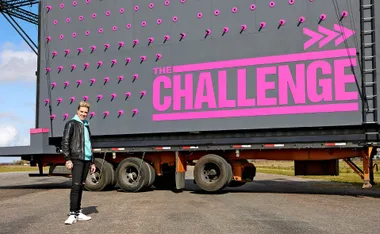 "Person standing in front of a large wall with pink pegs and 'The Challenge' text, outdoors."