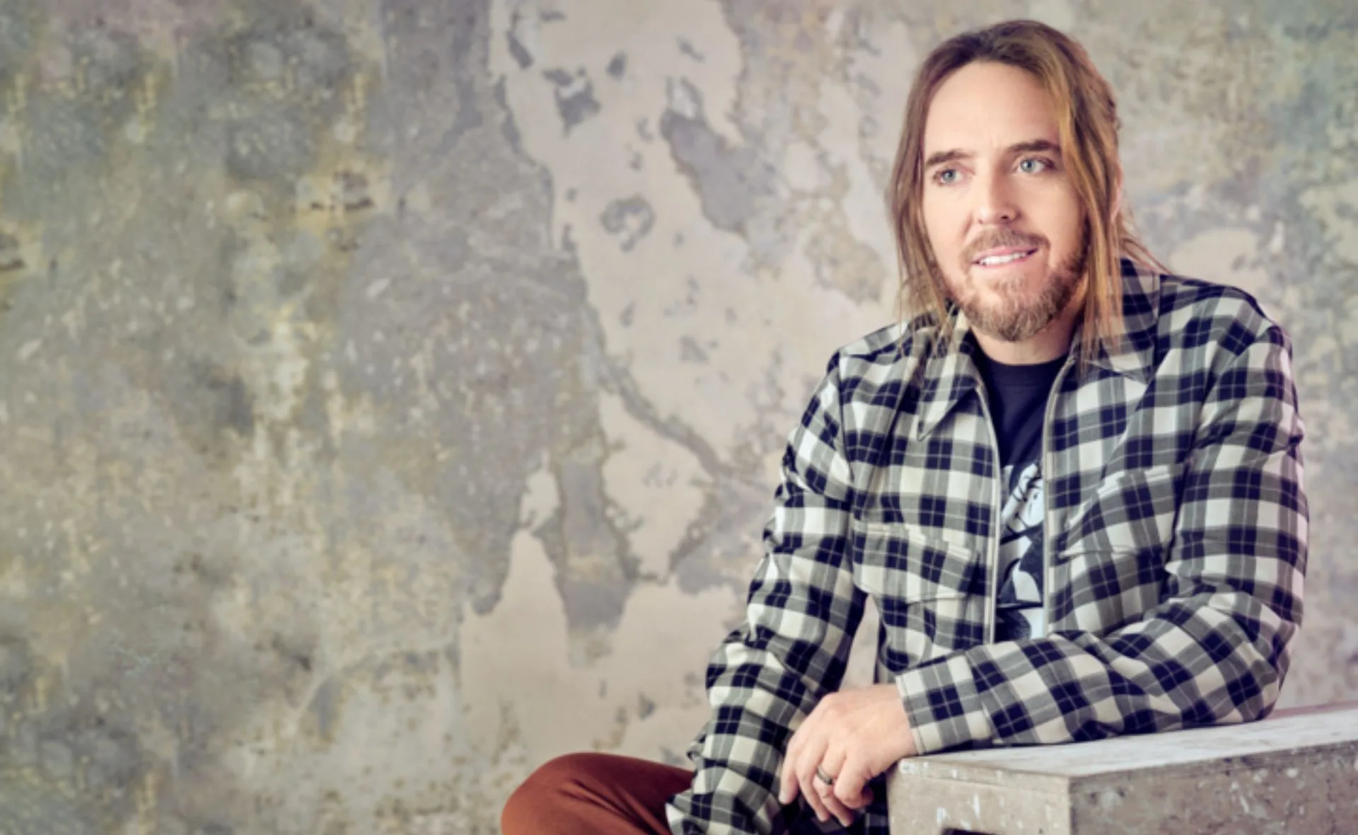 Man in plaid shirt sits against a textured wall, smiling softly.