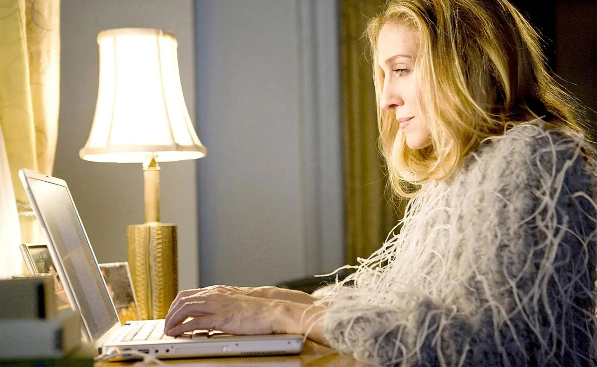 A woman in a feathered outfit using a laptop beside a lamp, in a cozy room setting.