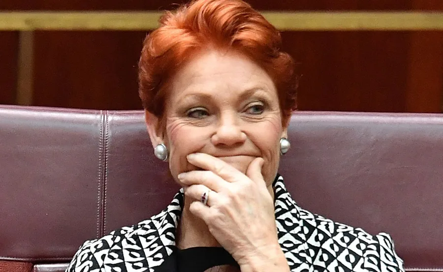 Woman with short red hair and pearl earrings smiling, wearing a patterned jacket, hand touching her chin.