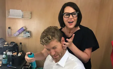 Two people in a dressing room; one is styling the other's hair with a spray can, both smiling and talking.
