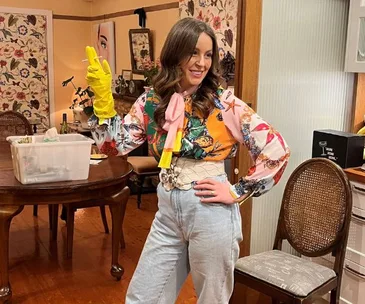 Woman in colorful blouse and jeans poses confidently in a living room, wearing a single yellow cleaning glove.