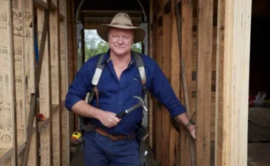 Man in construction attire with a hat, standing in a framed house, holding a hammer.