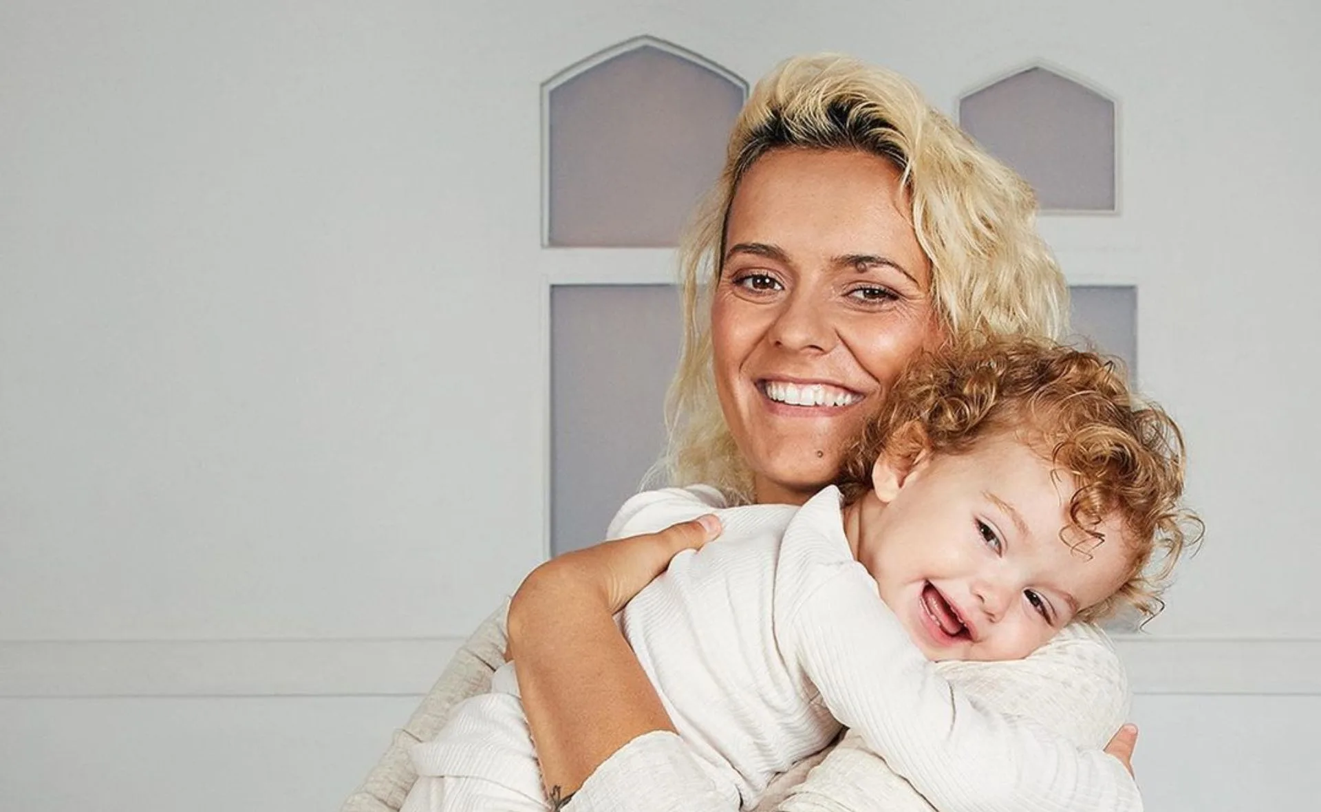 A person with blonde hair smiling, holding a joyful child with curly hair, both in light clothing, against a simple background.