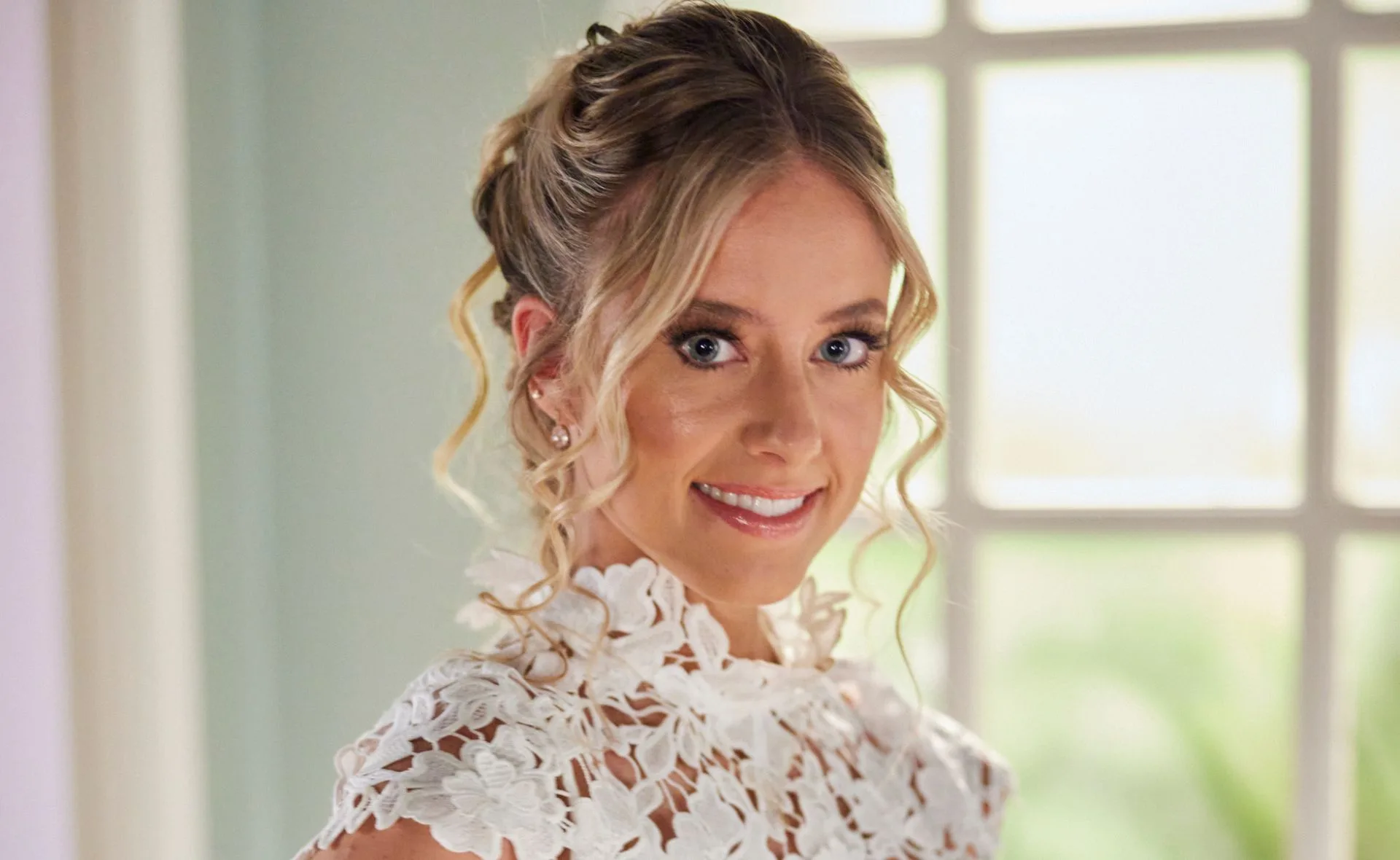 Woman smiling in a white floral lace dress, curly updo, indoors, bright background.