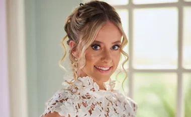 Woman smiling in a white floral lace dress, curly updo, indoors, bright background.