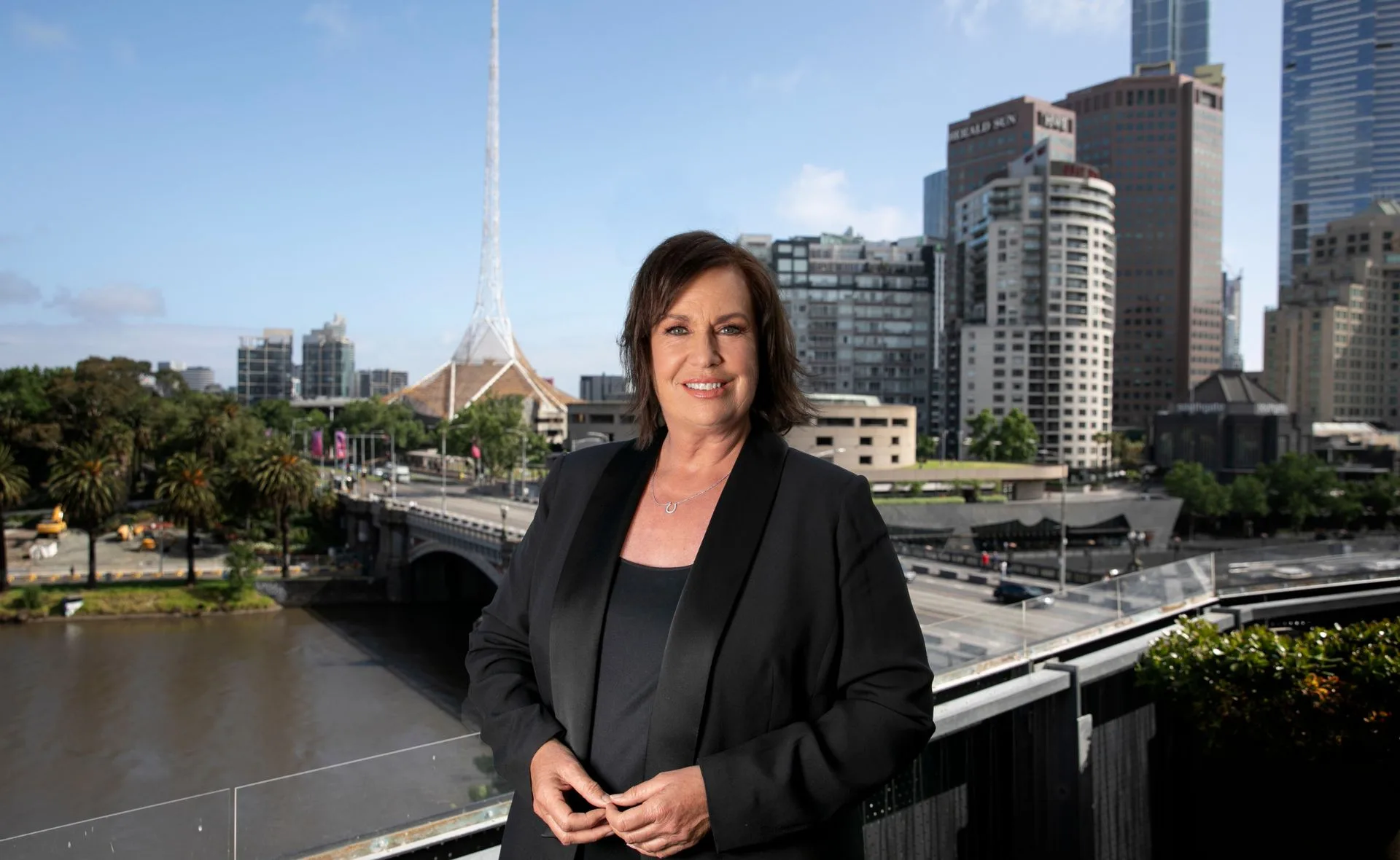 A person in a dark suit stands on a veranda with a cityscape, including modern buildings and a tower, in the background.