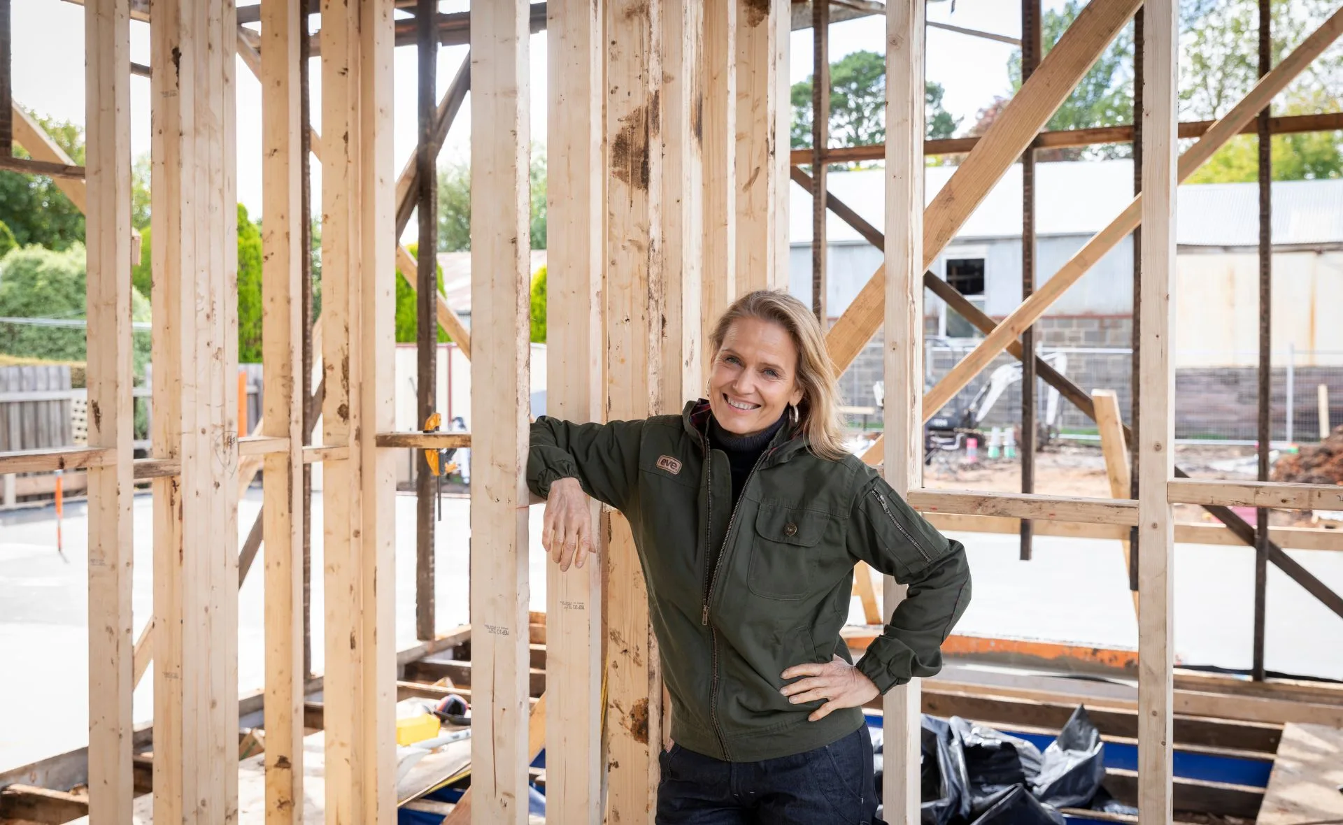 Woman in a green jacket leaning on wooden beams at a construction site, smiling.