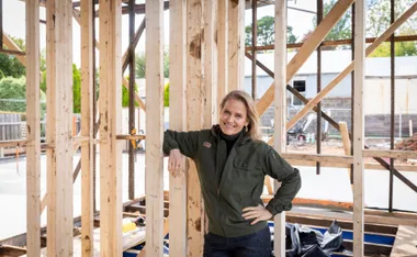 Woman in a green jacket leaning on wooden beams at a construction site, smiling.