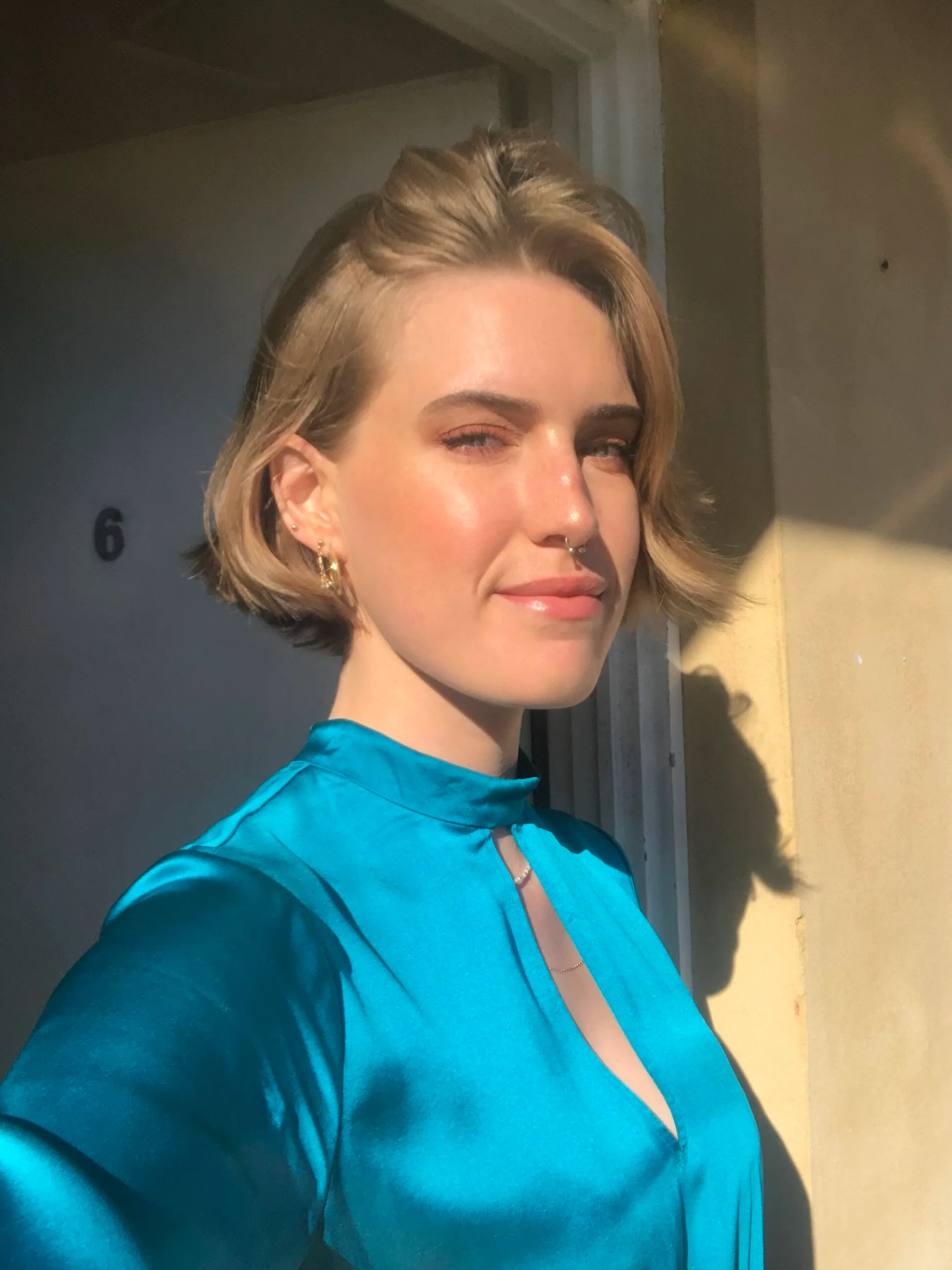 A person with short hair wearing a teal top stands in sunlight, casting a shadow against a wall numbered "6".