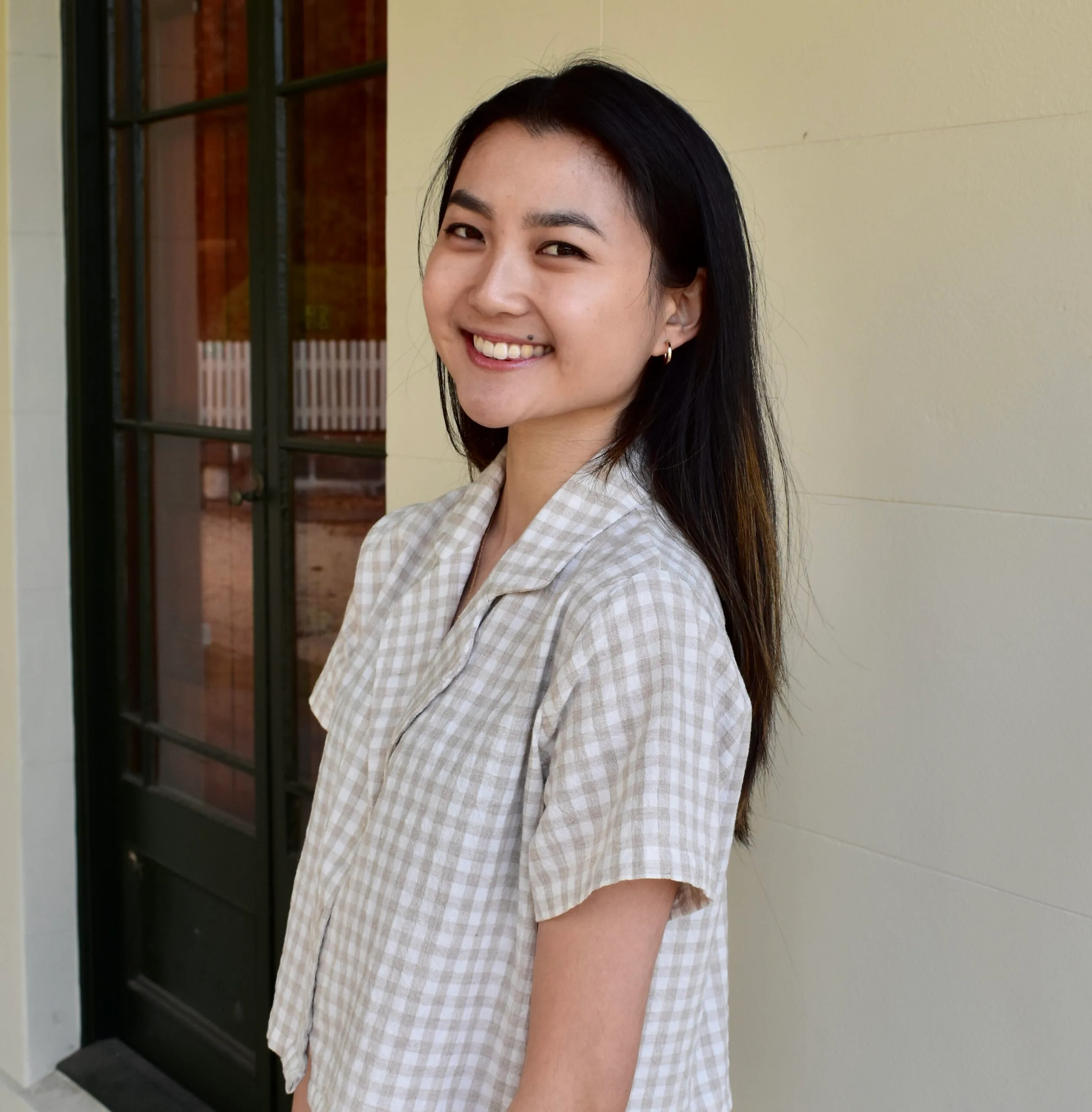 A person smiling, standing sideways, wearing a checkered shirt, with a window and cream wall in the background.