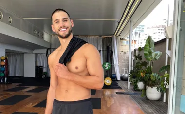 Man standing shirtless in a gym, holding a black shirt over his shoulder and smiling, surrounded by plants and gym equipment.