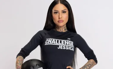 "Person with long dark hair, tattoos, wearing a 'The Challenge Jessica' shirt, holding a black helmet, poses confidently."