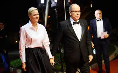 Couple walking on a red carpet, he in a tuxedo and she in a white shirt and black skirt, under event lights.