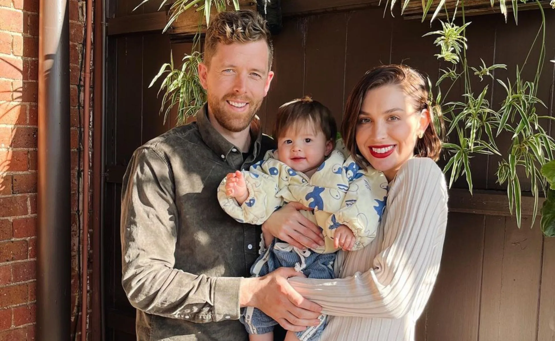 A family of three standing together, smiling, with the parents holding a baby in a patterned outfit, against a brick wall.