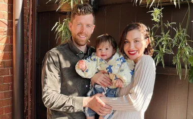 A family of three standing together, smiling, with the parents holding a baby in a patterned outfit, against a brick wall.