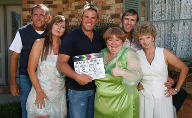 Cast members from "Kath & Kim" smiling with a clapperboard outside a brick house.