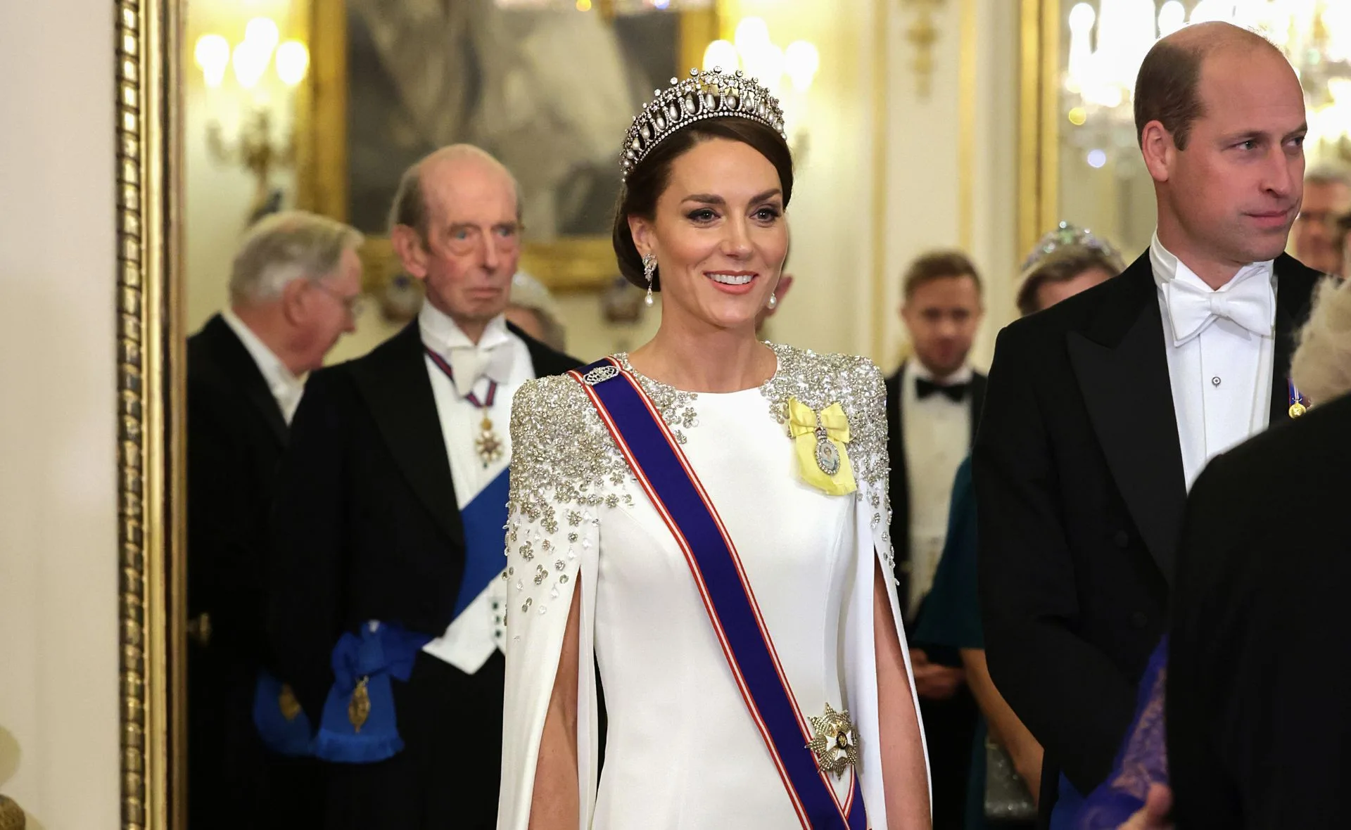 "Person in white dress and tiara smiling, with others in formal attire at an elegant event."