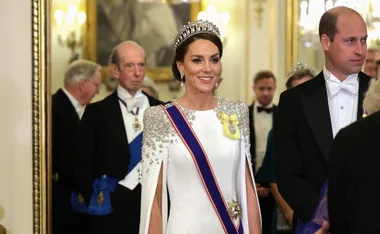 "Person in white dress and tiara smiling, with others in formal attire at an elegant event."